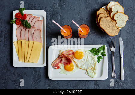 Englisches Frühstück mit Rührei und Speck, Karottensaft, Brot, Aufschnitt, Käse und Schinken auf einem dunklen Stein Hintergrund Stockfoto