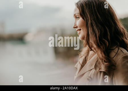Close up Portrait von freundlich charmante brünette Frau mit langen lockigen Haar im braunen Mantel an Straße mit goldenen Blätter, Herbst Stockfoto