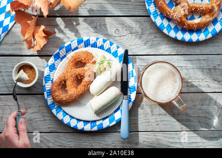 Oktoberfest traditionelles Essen und Bier. Hand, die Gabel mit Weißwurst, Brezeln auf Papptellern. Flach auf Holztisch mit blauen und weißen Pa Stockfoto