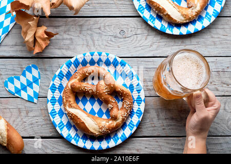 Oktoberfest traditionelles Essen und Bier. Hand, Bierkrug, Brezeln auf Papptellern. Flach auf Holztisch mit blauen und weißen Papier Bayerische de Stockfoto