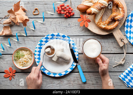 Oktoberfest traditionelles Essen und Bier. Hände halten Bierkrug und Gabel mit Weißwurst, Brezeln auf Papptellern. Flach auf Holztisch mit Blu Stockfoto