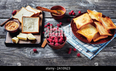 Toast von Weizen Brot auf dem Teller mit zerlassener Butter in eine Schüssel geben und frische Himbeeren auf einem Holztisch, Zutaten zum Kochen Sandwiches mit Erdnussbutter bu Stockfoto