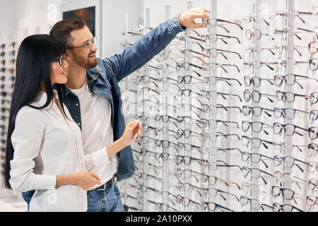 Bärtige handsoem lächelnden Mann Gläser aus dem Regal zu nehmen, um sie auszuprobieren, Nahaufnahme, Seite, Ansicht Foto. Kopieren Sie Platz Stockfoto