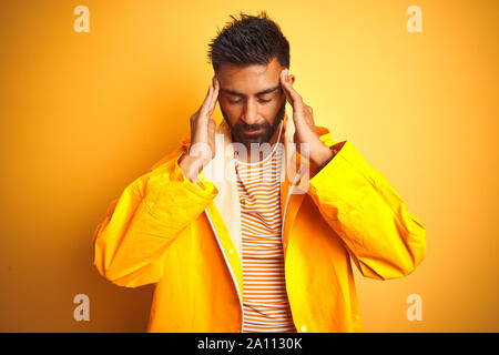 Junge indische Mann mit regenmantel stehend über isolierte gelber Hintergrund mit der Hand auf den Kopf für Schmerzen im Kopf, weil Stress. Migräne leiden. Stockfoto