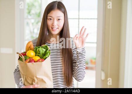 Schöne asiatische Frau mit papiertüte von frischen Lebensmitteln zu tun ok Zeichen mit den Fingern, ausgezeichnete Symbol Stockfoto
