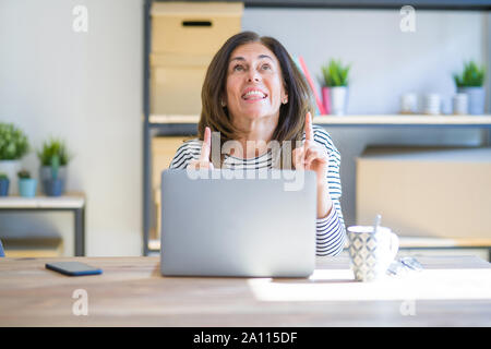 Mittleren Alters ältere Frau am Tisch zu Hause sitzen Arbeiten mittels Computer Laptop erstaunt und überrascht mit der Suche und zeigt mit Fingern und Heben Stockfoto