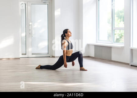 Junge schöne sportliche Frau mit schwarzen Sportswear sport Übungen, ausfallschritte, in der Nähe der Fenster in der Turnhalle, Stretching und Beinen stehend auf Asche Stockfoto