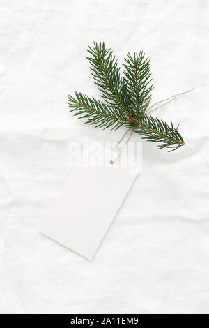 Close-up leeres weißes Papier Geschenkanhänger mit Seil und Grüne Tanne Zweig auf weißem Leinen Tischdecke Hintergrund. Preis nmock-up-Szene. Weihnachten gesundes Stockfoto