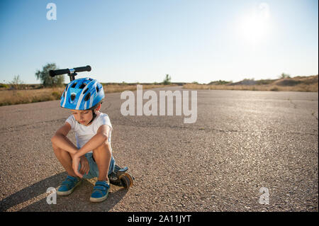 Traurigen kleinen Jungen in der Helm sitzt auf seinem Roller auf Asphalt leere Straße im Sommer heiß Sonnenuntergang in Kalifornien Stockfoto