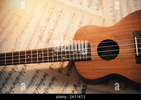 Eine kleine hölzerne Ukulele mit Nylonsaiten liegt auf verstreute Blätter der Noten, durch das Licht leuchtet Stockfoto