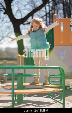 Cute curly blonde Mädchen in Blau aufgeblasen Weste freut und spielt auf dem Spielplatz im Stadtpark. Stockfoto