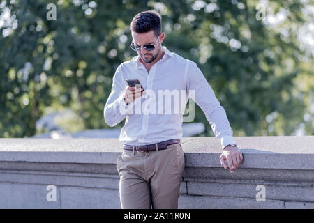 Ein hübscher junger Geschäftsmann auf der Brücke stehen und mit seinem Smartphone an einem sonnigen Tag. Stockfoto