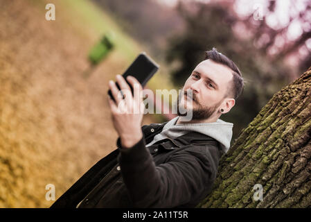 Ein hübscher junger hipster Mann lehnte sich an einen Baum und ein selfie im Park. Stockfoto