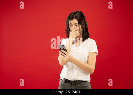 Der Kaukasischen junge Frau Brustbild auf Roten studio Hintergrund. Schöne weibliche Modell im weißen Hemd. Konzept der menschlichen Gefühle, Mimik. Kauft online, Bezahlung Rechnung, schaut entsetzt. Stockfoto