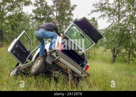 Der Mann an der Spitze der klemmt SUV, versuchen Sie, den eingeklemmten Auto aus der Grube zu erhalten.'s Rad klemmt. Weißes Auto auf einem grünen Wald Hintergrund, Park Stockfoto