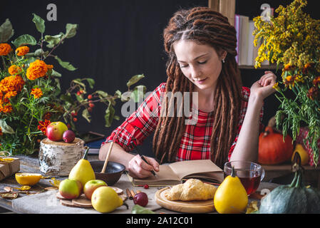 Schöne junge Frau schreibt Herbst Träume in einem Notebook. Fallen Tisch mit Blumen. Das Konzept der Herbst romantische Stimmung Stockfoto