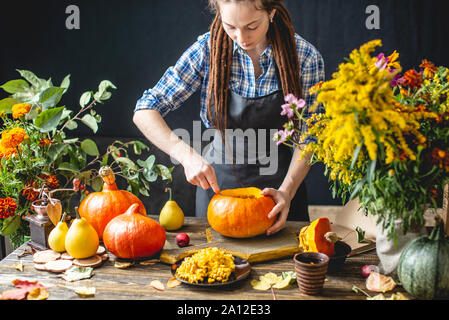 Eine Frau Koch reinigt eine orange Kürbis zum Schnitzen für die Halloween Urlaub. Konzept herbst Essen in einer gemütlichen Küche aus dunklem mit gelben Blumen Stockfoto