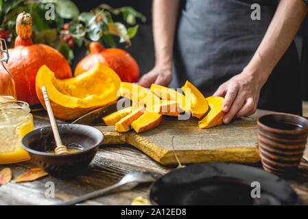 Eine Frau Koch schneidet ein orange Kürbis mit einem Messer in Scheiben zum Backen auf einem Holztisch. Konzept herbst Essen in einer gemütlichen dunklen Küche mit gelben Fluss Stockfoto