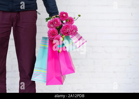 Stilvolle Mann mit Blumen und rosa, blau Einkaufstaschen auf Stein Hintergrund. Tag der Frau. Valentinstag. Bereit für die Geburtstagsfeier oder romantisches Date. Stockfoto