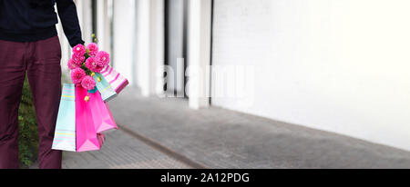 Stilvolle Mann mit Blumen und rosa, blau Einkaufstaschen auf Stein Hintergrund. Tag der Frau. Valentinstag. Bereit für die Geburtstagsfeier oder romantisches Date. Stockfoto