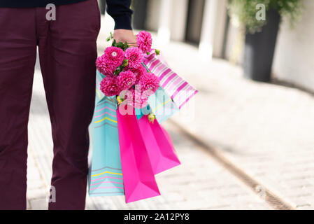 Rosa Blumen in männlichen Händen und Einkaufstaschen auf Stein Hintergrund. Geburtstag, Mutter, Frauen, Hochzeit Konzept. Bündel Papier Geschenkbeutel. Stockfoto