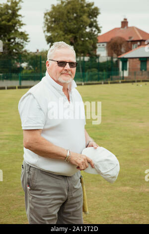 Ein Porträt von einem älteren Mann grinsend zu einem Bowling Green. Er trägt eine Sonnenbrille und eine weiße Kappe. Stockfoto