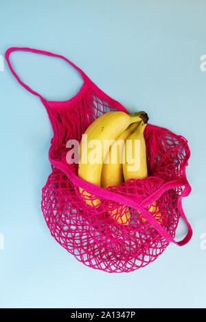 Drei gelbe reife Bananen in einem Fuchsia string Beutel auf Pastell-blaue Hintergrund. Wiederverwendbaren Netzbeutel oder Mesh Shopper. Flach. Ansicht von oben. Stockfoto