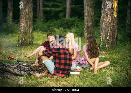 Gerne Freunde sprechen um Lagerfeuer. Kerle kochen Würstchen für Abendessen im Freien. Übernachtung in Zelten im Wald, Picknick im Wald. Stockfoto