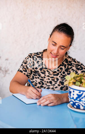 Frau schreiben Sie Notizen in Ihr Notizbuch auf Blau Schreibtisch außerhalb, tragen Animal-print Shirt. Stockfoto