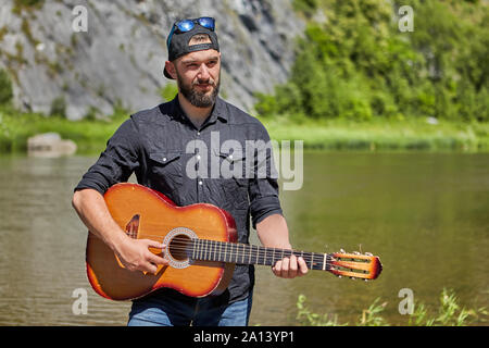 Eine 23 Jahre alte bärtige Mann spielt eine akustische Gitarre im Freien gegen einen wilden Fluss. Ein junger Mann von einem kaukasischen Ethnie verwendet ein Musikinstrument in der Na Stockfoto