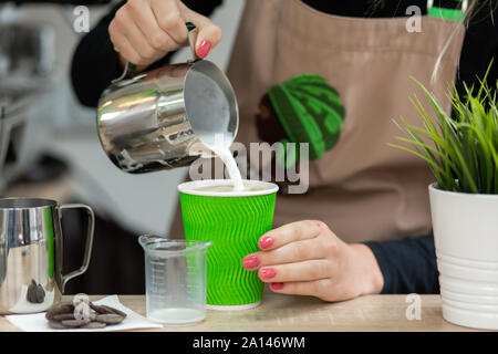 Barista in eine Schürze gießt Milch in ein Grün umweltfreundliche Pappbecher. Barista arbeiten in einem Coffee Shop. Stockfoto