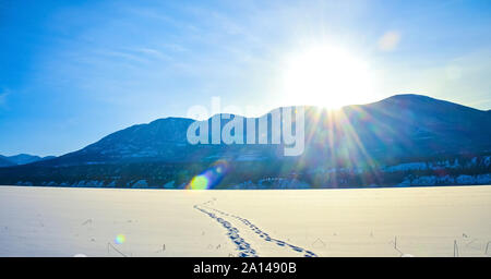 Fuß Drucke im Schnee über einen zugefrorenen See mit Sun flare und schneebedeckten Bergen im Hintergrund. Ein Winter Berge in British Colum Stockfoto