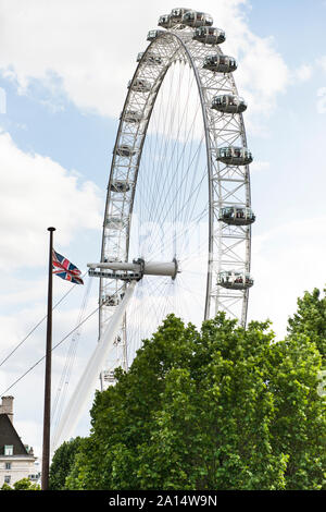 Blick auf das London Eye mit der britischen Flagge entrollt und grüne Bäume im Vordergrund. Schönen, sonnigen Tag und flauschige Wolken. Kopieren Sie Platz. Stockfoto