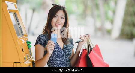 Junge asiatische Frau mit Kreditkarte und Shopping Bag nach Wegnahme von Bargeld aus Geldautomaten Stockfoto