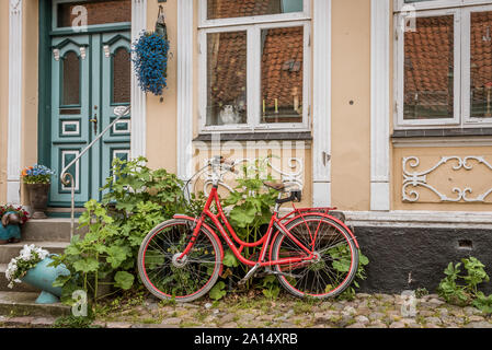 Ein rot Retro Fahrrad auf Kopfsteinpflaster, schiefen aganst eine Wand mit hollyhochs in ÆRØSKØBING, Dänemark, 13. Juli 2019 Stockfoto