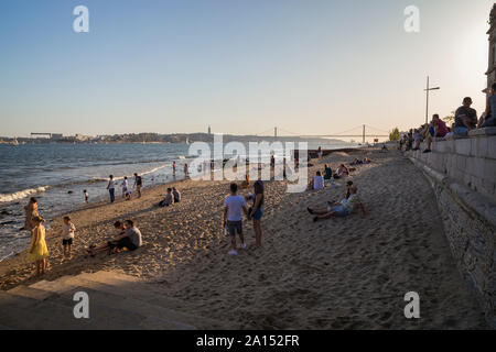 Menschen entspannen an einem kleinen Strand in der Innenstadt von Lissabon, Portugal, an einem sonnigen Nachmittag. Brücke "25 de Abril" und Cristo Rei Monument sind im Hintergrund. Stockfoto