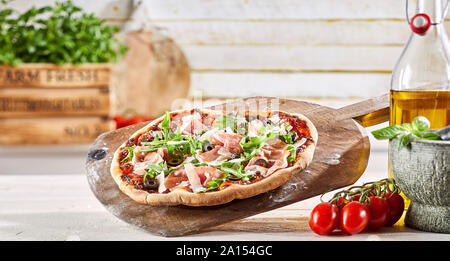 Dünne Kruste italienische Pizza mit Schinken, frischen Rucola Blätter, Parmesan Flocken und Oliven auf einem hölzernen Paddel mit Zutaten in eine Pizzeria Stockfoto