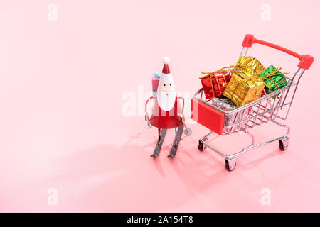 Christmas shopping Konzept, mini Rot Warenkorb Trolley mit Santa Claus Spielzeug und Geschenkbox auf rosa Hintergrund, leere Kopie Platz, in der Nähe Stockfoto