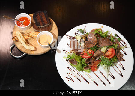 Salat mit Rindfleisch, Zunge mit Gemüse und Kräutern, gekleidet mit Soße auf einer weißen Platte. Gebratene Rinderleber mit Pilzen, Gemüse und Gewürzen. Stockfoto