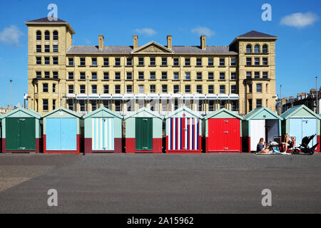 Brighton, East Sussex, England, UK, 13. September 2019: eine Linie von bunten Badekabinen mit einer Familie sitzt außerhalb einer von Ihnen, hinter ist eine große b Stockfoto