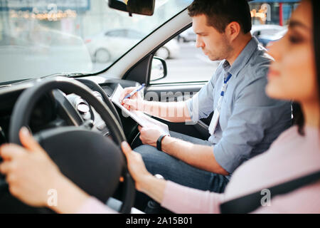 Männliche auto Kursleiter nimmt Untersuchung in der jungen Frau. Modell Auto. Die Hände am Lenkrad festhalten. Männliche Kursleiter Schreiben auf Papier. Road Trip Stockfoto