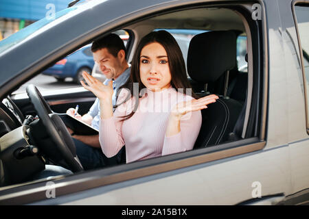 Männliche auto Kursleiter nimmt Untersuchung in der jungen Frau. Sie sitzen auf der Fahrer und Blick auf Kamera mit Verwirrung. Betonte frau Hände schütteln. Kursleiter si Stockfoto