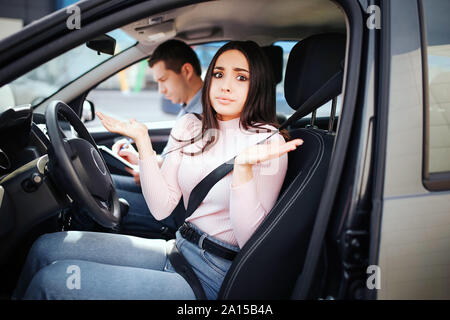 Männliche auto Kursleiter nimmt Untersuchung in der jungen Frau. Zusammen Sitzen im Auto. Verwirrt Frau Blick auf Kamera und Welle mit Händen. Besetzt Kursleiter zu schreiben. Exa Stockfoto