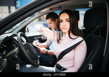 Männliche auto Kursleiter nimmt Untersuchung in der jungen Frau. Schöne brünette Modell sieht traurig auf Kamera. Fehlerhafte Prüfungen. Kursleiter neben ihr und weißen Sitzen Stockfoto