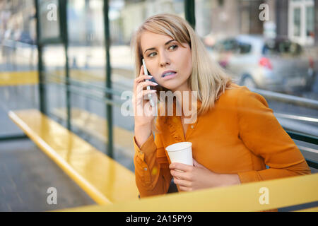 Junge Frau mit Smartphone und Kaffee zum Mitnehmen warten auf den Bus an der Haltestelle Stockfoto