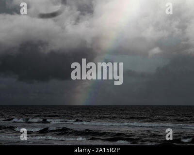 Meereslandschaft in monochromer Teilfarbe, Ein farbenfroher Regenbogen reicht vom Himmel bis zum Meer, dramatisches Wetter, stimmungsvoller dunkler stürmischer grauer Himmel Stockfoto