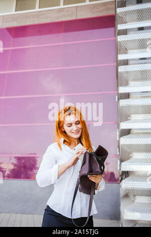 Lächelnd Business woman putting Smartphone in Handtasche vor einem modernen kommerziellen Gebäude Stockfoto