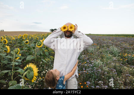 Verspielter Mann, die seine Augen mit Sonnenblumen auf einem Feld mit ihm Tochter umarmen Stockfoto