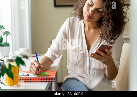 Junge Frau mit Handy und Notizen am Schreibtisch zu Hause Stockfoto
