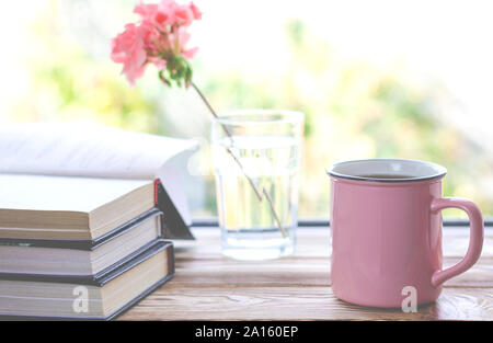 Eine rosa Tasse mit Kaffee steht auf einem hölzernen Fensterbank neben Büchern und ein Glas, in dem steht ein rosa Geranien Blume Stockfoto